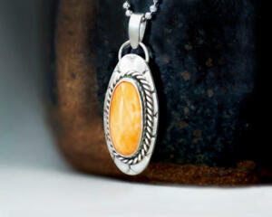 3/4 left view hanging on black pot, oval orange stone bezel set accented with twisted wire and a stamped base, diamond cut chain